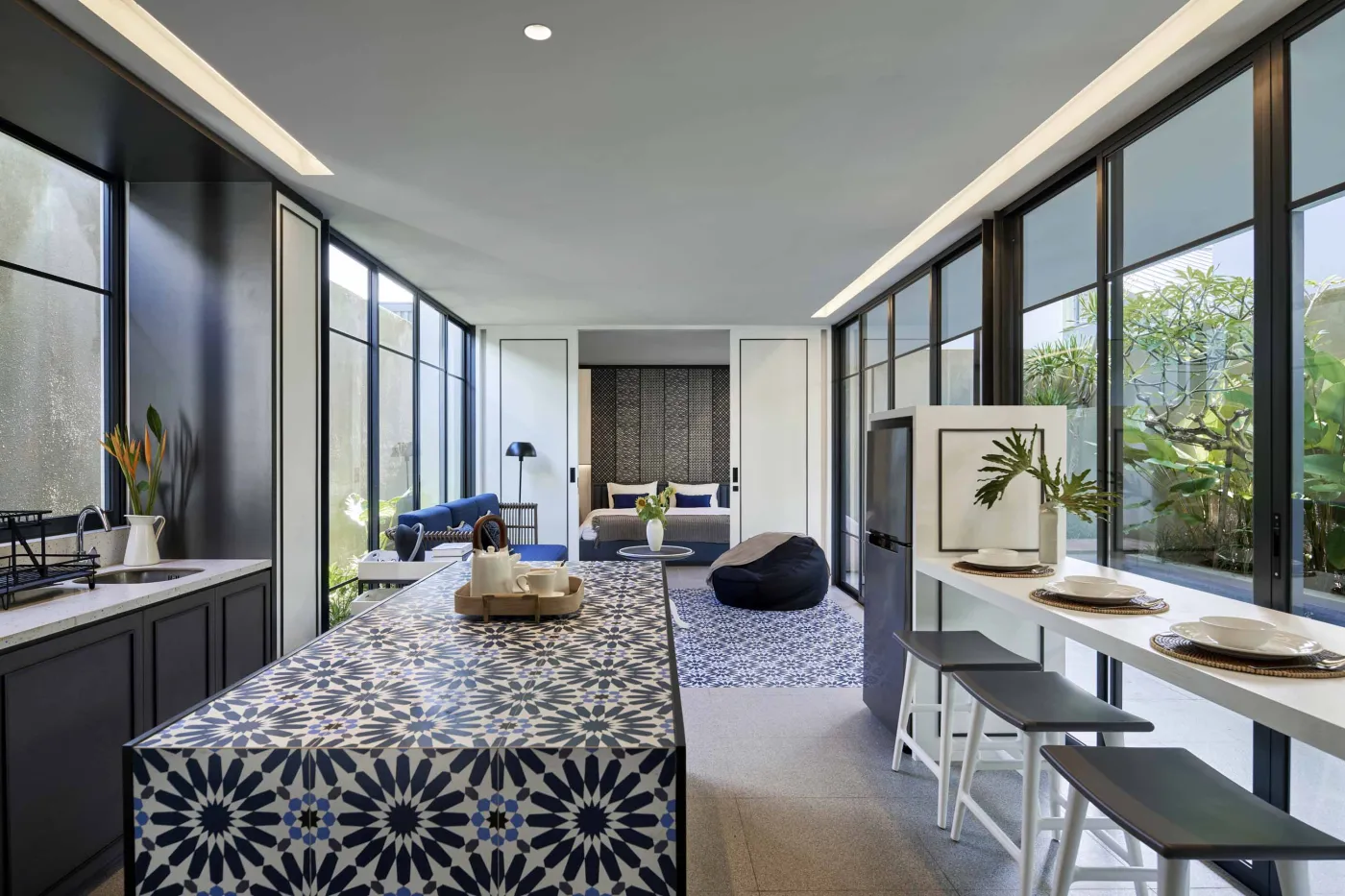 Modern open-plan room with blue mosaic tiled kitchen island, bar stools, blue sofa, bed, and large windows overlooking tropical garden. - Bali Villas