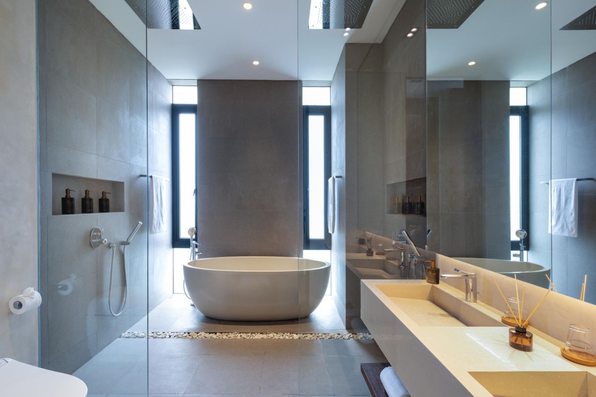 Modern luxury bathroom with freestanding white bathtub, gray stone walls, glass shower, and minimalist vanity. - Bali Villas