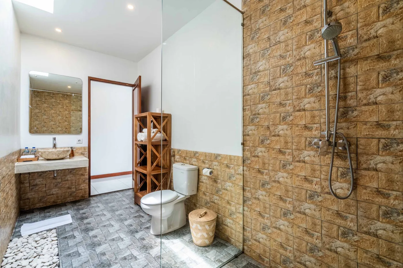 Modern tropical bathroom featuring brick-tiled shower, wooden vanity sink, toilet, and woven accents. - Bali Villas