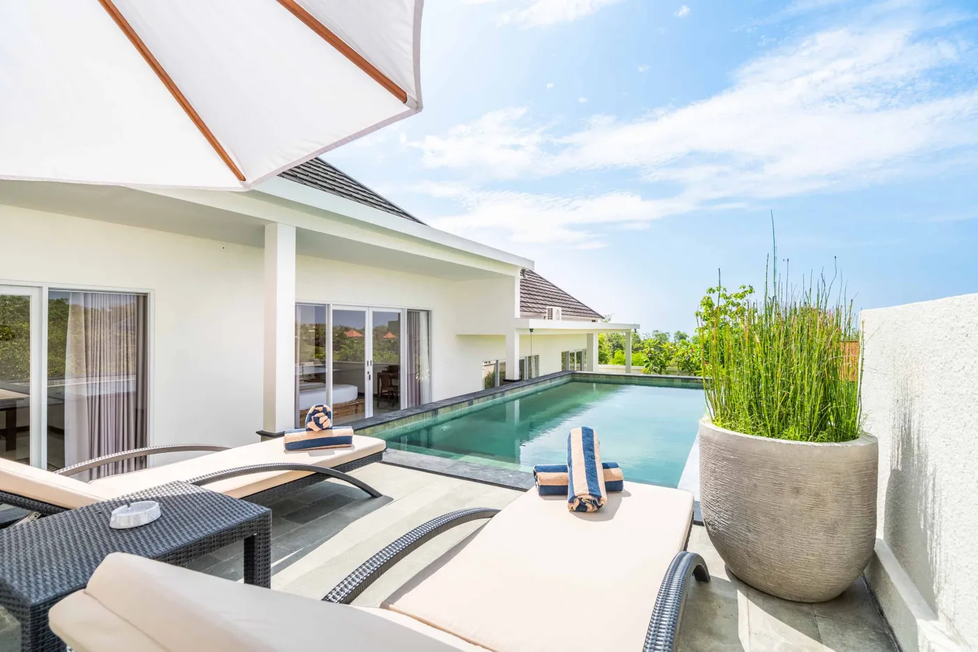 Luxurious infinity poolside at a modern white Bali villa, with lounge chairs, towels, umbrella, and tropical greenery under a sunny blue sky. - Bali Villas