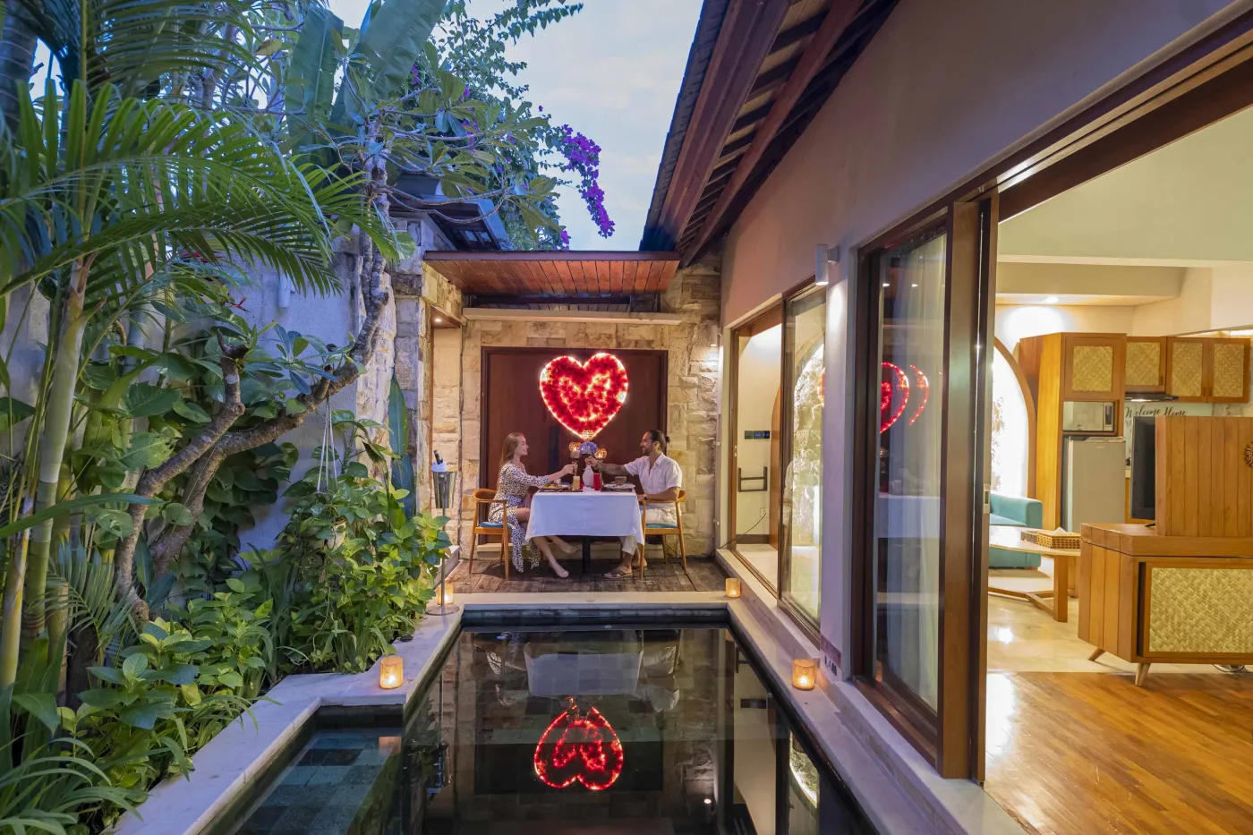 Romantic couple enjoying candlelit dinner by neon "LOVE" heart and reflecting pool in lush Balinese villa garden. - Bali Villas