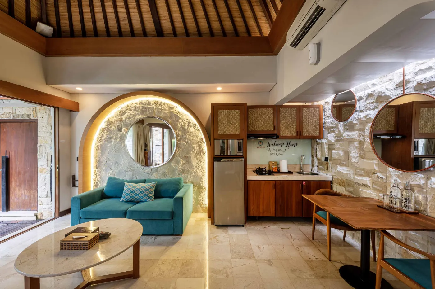 Cozy Balinese villa interior with thatched ceiling, turquoise sofa, wooden kitchenette, arched stone doorway, and round mirrors. - Bali Villas