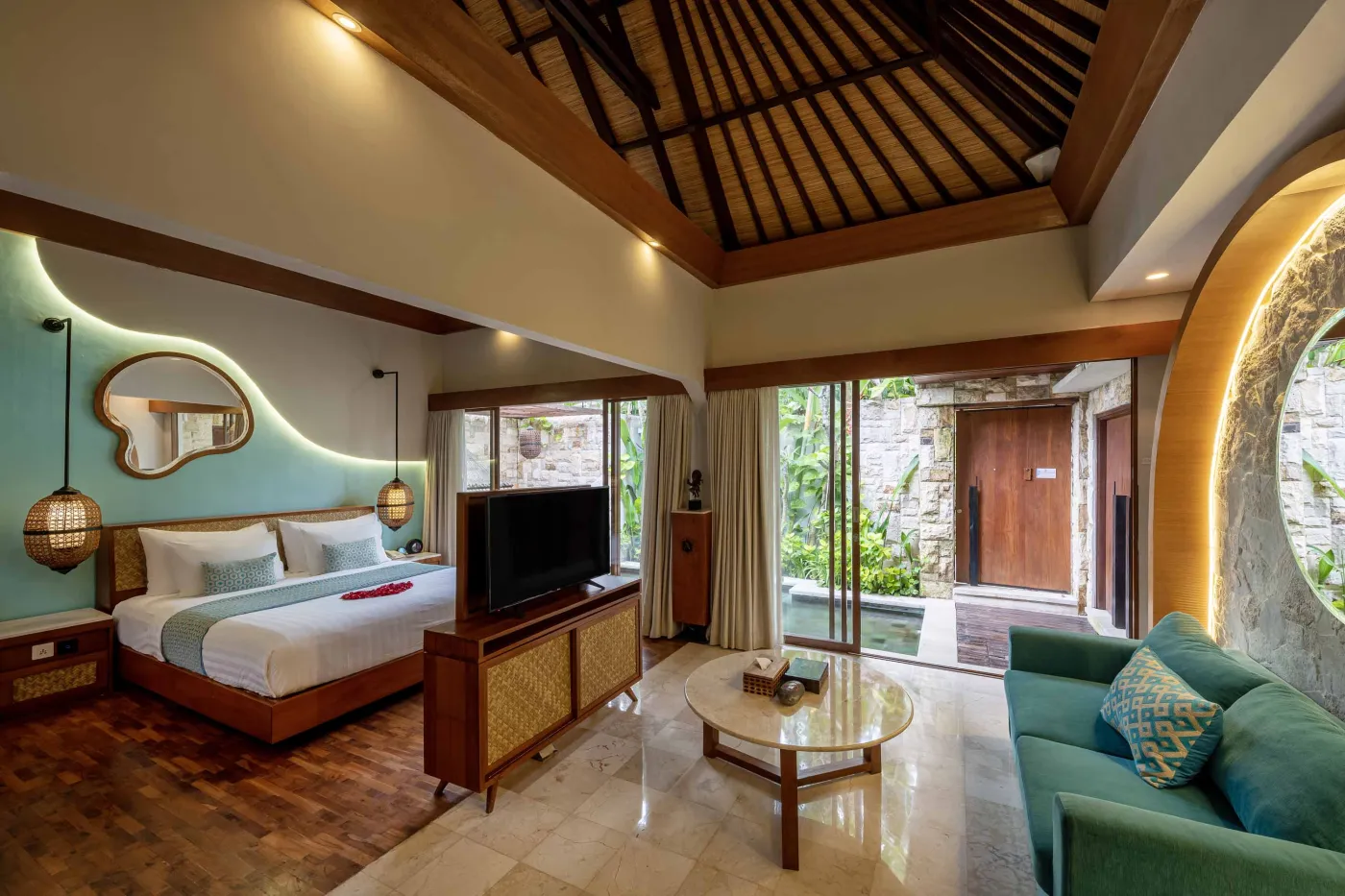 Cozy Balinese villa interior with thatched ceiling, turquoise sofa, wooden kitchenette, arched stone doorway, and round mirrors. - Bali Villas