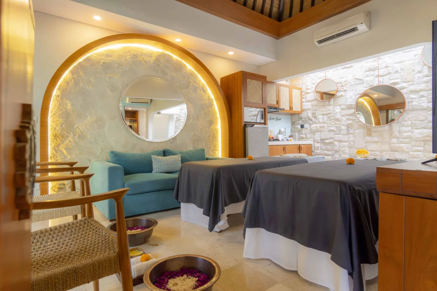 Cozy Balinese villa interior with thatched ceiling, turquoise sofa, wooden kitchenette, arched stone doorway, and round mirrors. - Bali Villas