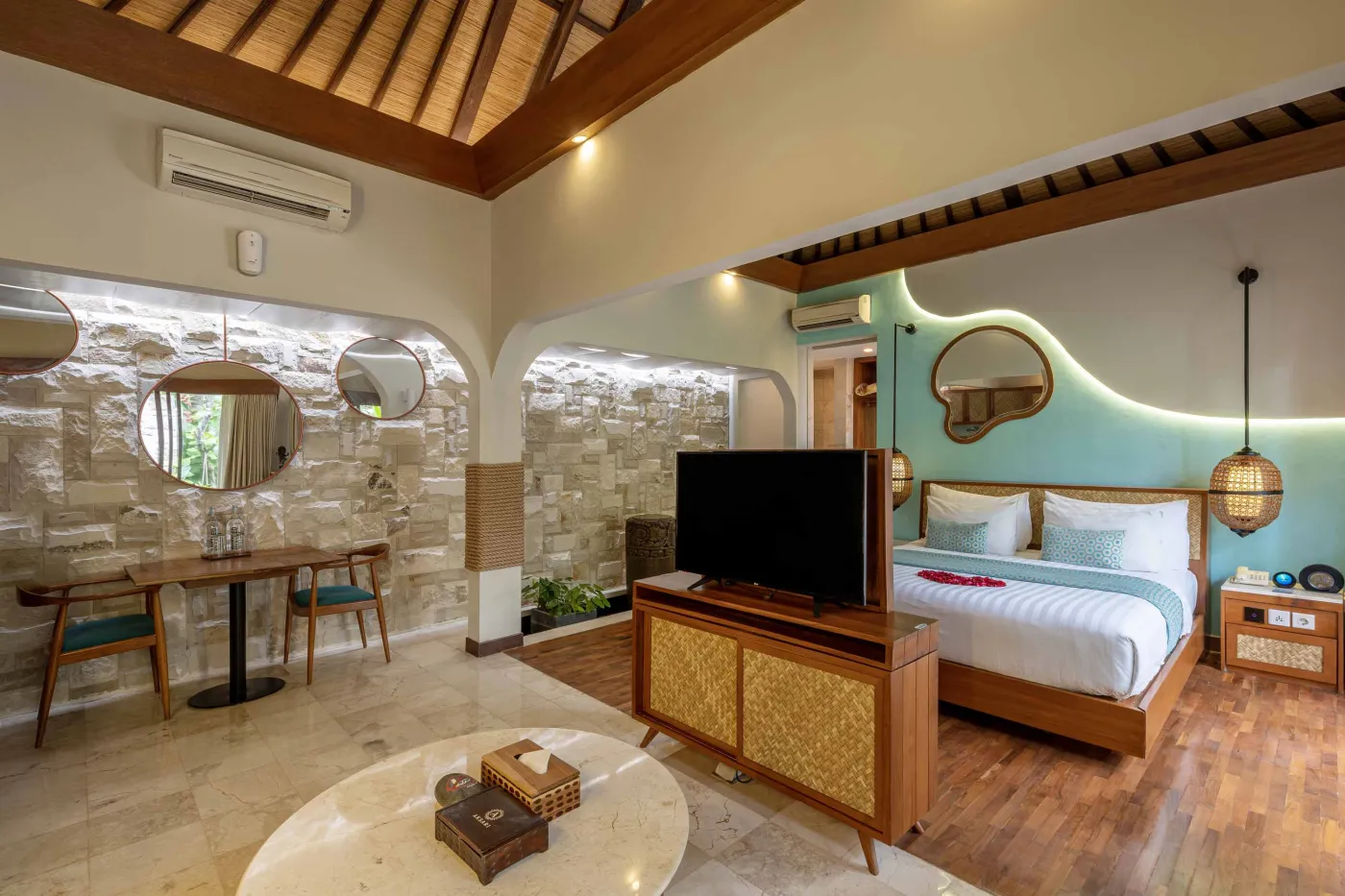 Cozy Balinese villa interior with thatched ceiling, turquoise sofa, wooden kitchenette, arched stone doorway, and round mirrors. - Bali Villas