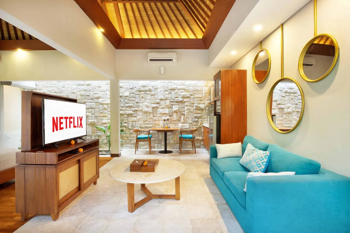 Cozy Balinese villa interior with thatched ceiling, turquoise sofa, wooden kitchenette, arched stone doorway, and round mirrors. - Bali Villas