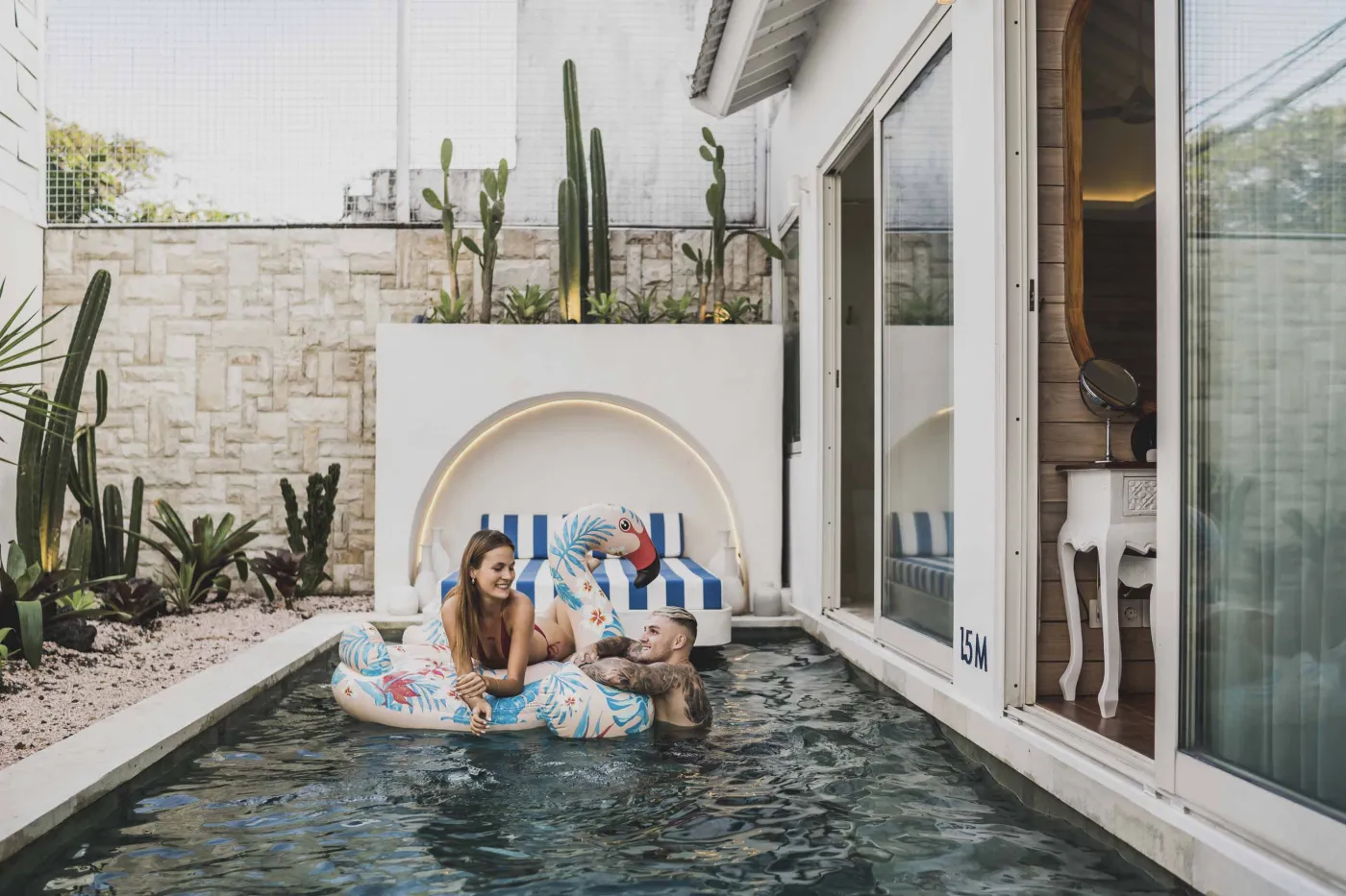 Modern Balinese-style reception lobby with wooden desk, blue armchairs, tropical plants, and glass doors to outdoor greenery. - Bali Villas