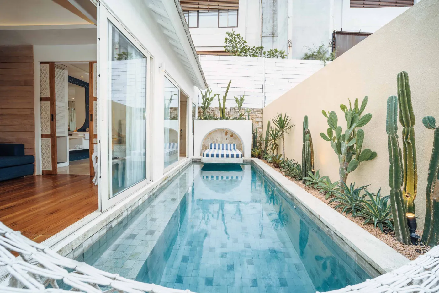 Modern Balinese-style reception lobby with wooden desk, blue armchairs, tropical plants, and glass doors to outdoor greenery. - Bali Villas