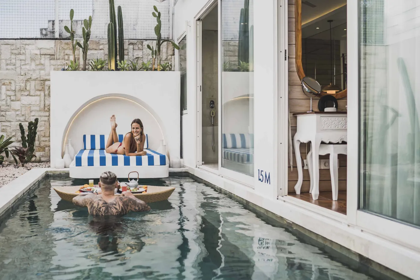 Modern Balinese-style reception lobby with wooden desk, blue armchairs, tropical plants, and glass doors to outdoor greenery. - Bali Villas