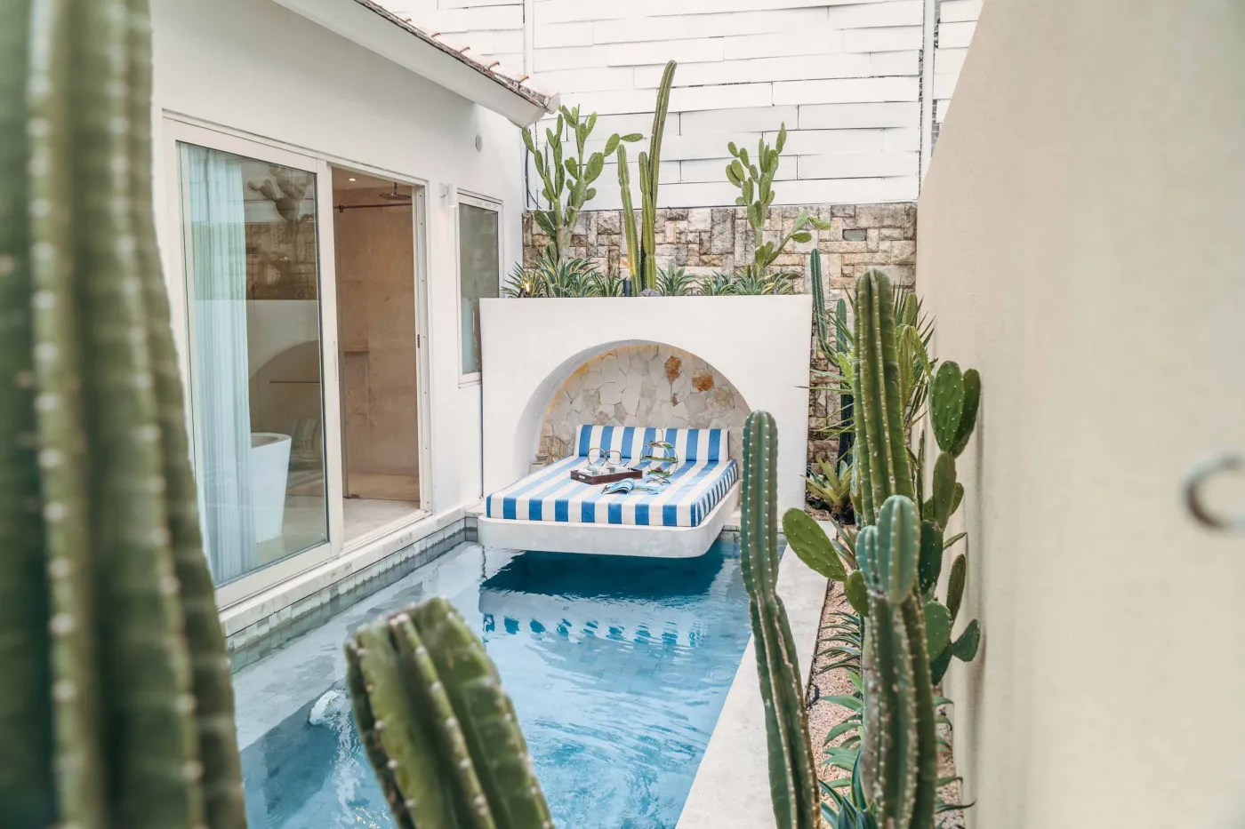Modern Balinese-style reception lobby with wooden desk, blue armchairs, tropical plants, and glass doors to outdoor greenery. - Bali Villas
