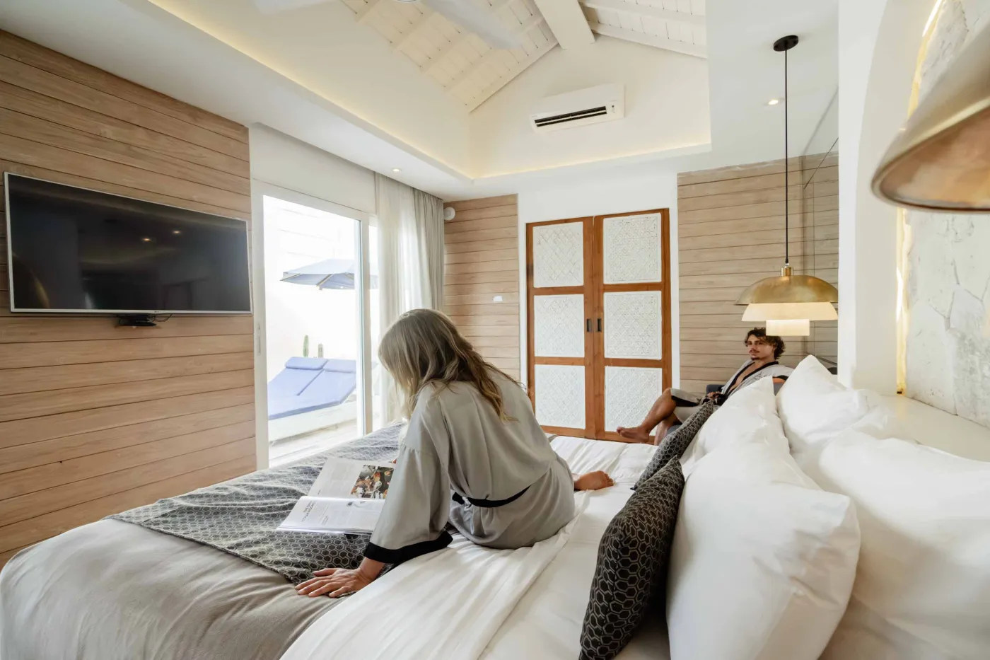 Modern Balinese-style reception lobby with wooden desk, blue armchairs, tropical plants, and glass doors to outdoor greenery. - Bali Villas