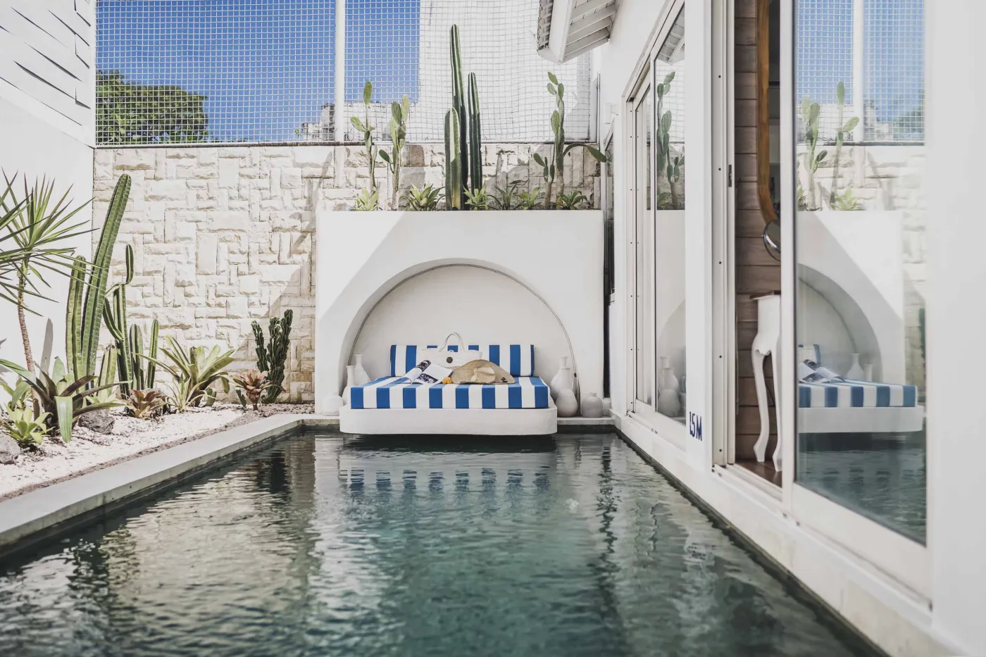 Modern Balinese-style reception lobby with wooden desk, blue armchairs, tropical plants, and glass doors to outdoor greenery. - Bali Villas