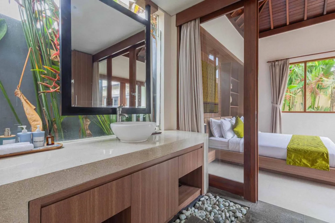 Modern Balinese villa interior: open kitchen with wooden cabinets, granite island, tiled backsplash, and dining table with pink orchids, overlooking a green balcony. - Bali Villas