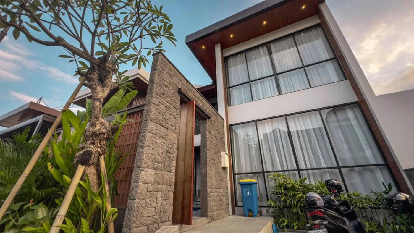 Modern Balinese villa interior: open kitchen with wooden cabinets, granite island, tiled backsplash, and dining table with pink orchids, overlooking a green balcony. - Bali Villas