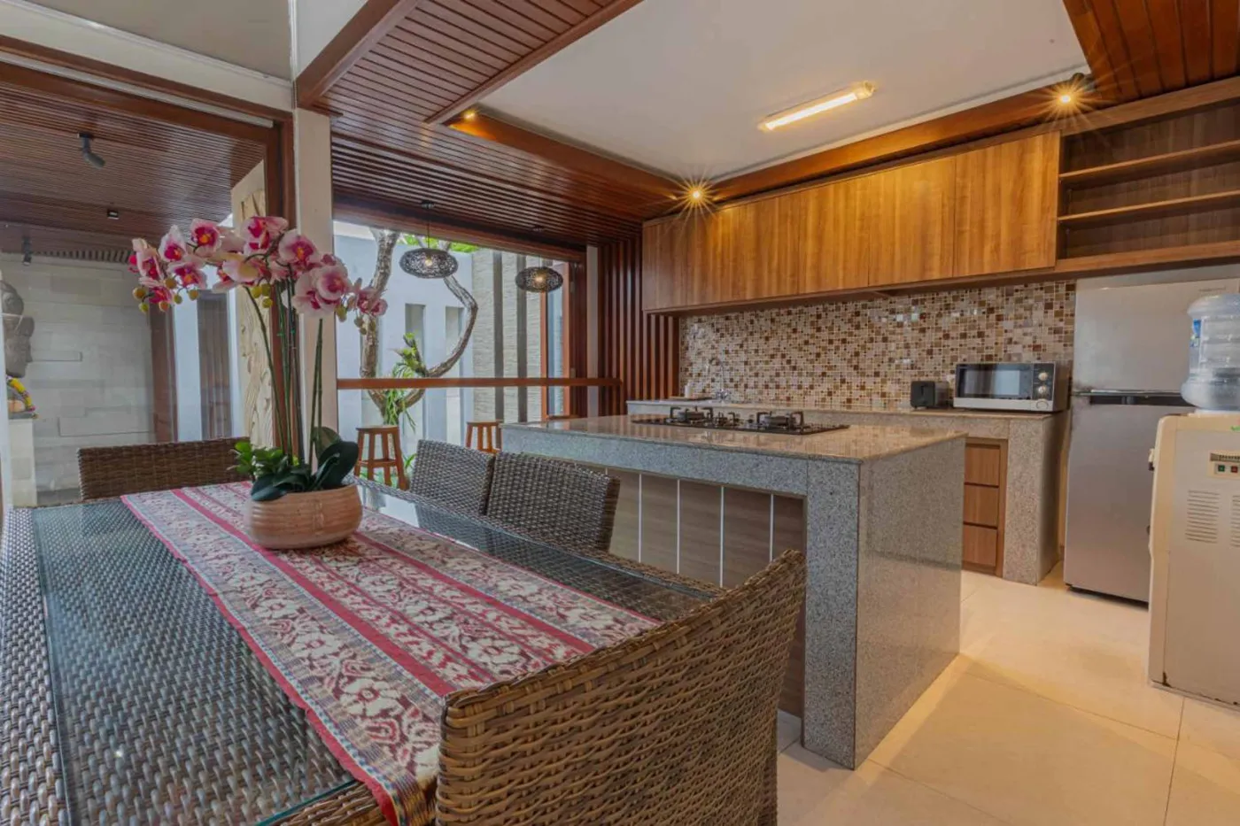 Modern Balinese villa interior: open kitchen with wooden cabinets, granite island, tiled backsplash, and dining table with pink orchids, overlooking a green balcony. - Bali Villas