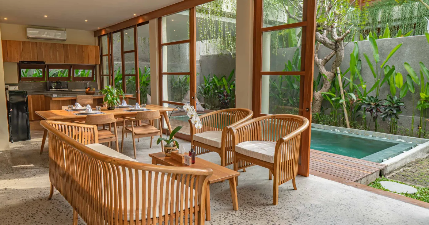 Spacious wooden kitchen and dining area in a tropical Balinese-style villa, featuring a set table with plates, glasses, vibrant floral centerpiece, and lush garden view through open sliding doors. - Bali Villas