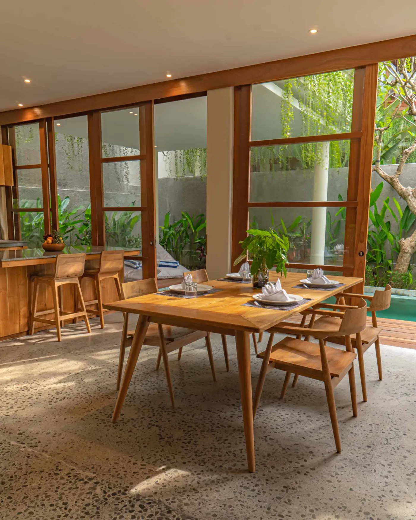 Spacious wooden kitchen and dining area in a tropical Balinese-style villa, featuring a set table with plates, glasses, vibrant floral centerpiece, and lush garden view through open sliding doors. - Bali Villas