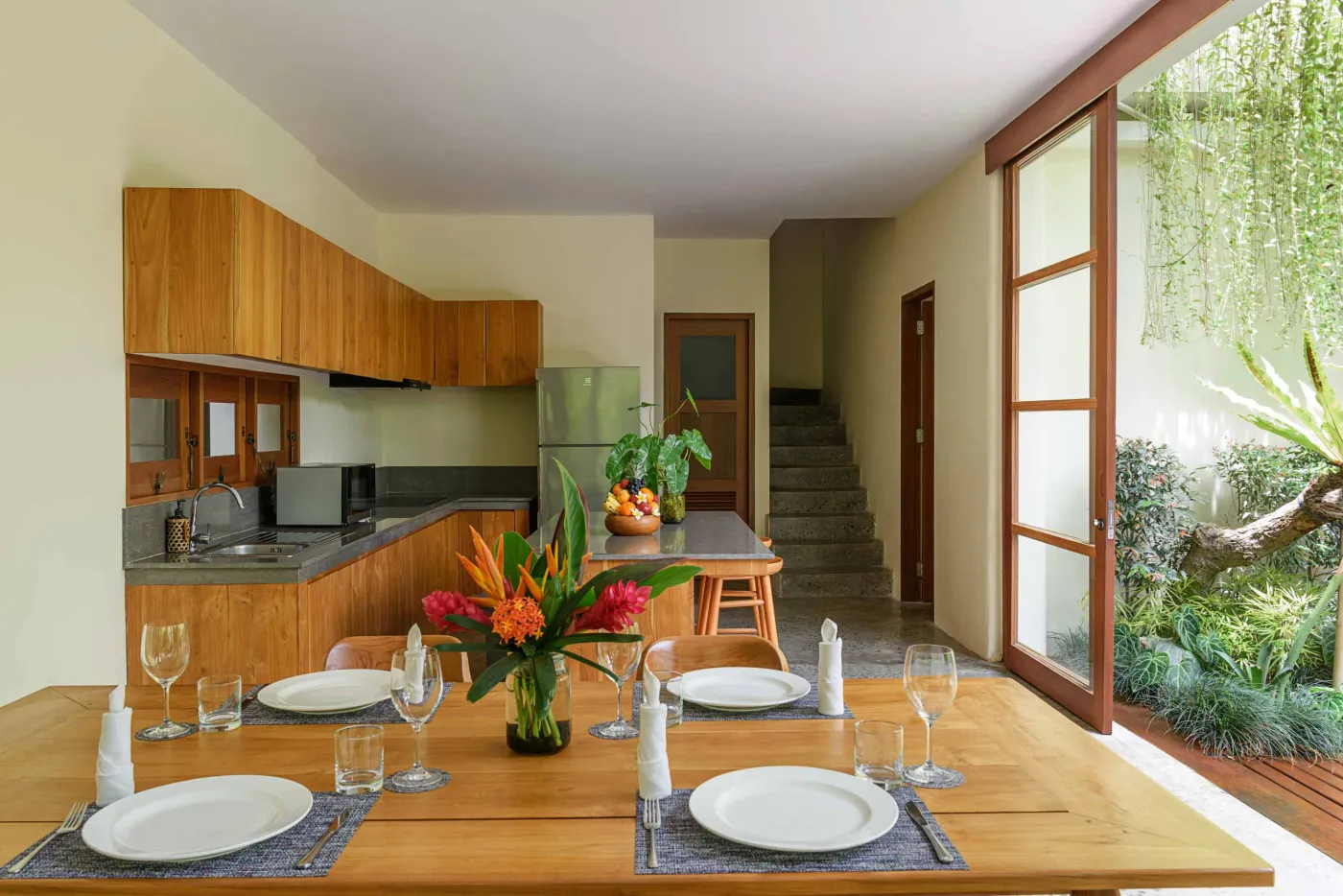 Spacious wooden kitchen and dining area in a tropical Balinese-style villa, featuring a set table with plates, glasses, vibrant floral centerpiece, and lush garden view through open sliding doors. - Bali Villas
