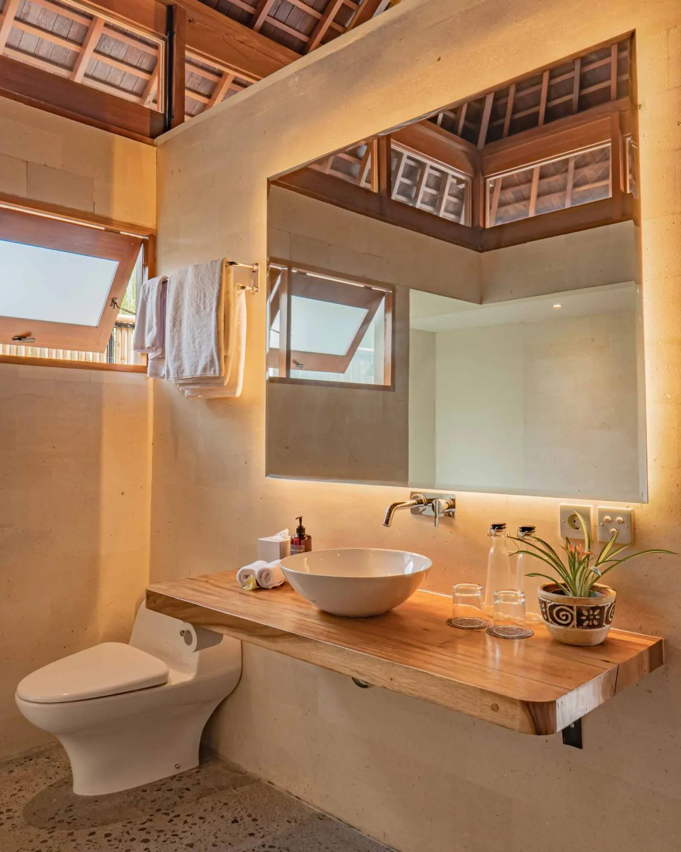 Spacious wooden kitchen and dining area in a tropical Balinese-style villa, featuring a set table with plates, glasses, vibrant floral centerpiece, and lush garden view through open sliding doors. - Bali Villas