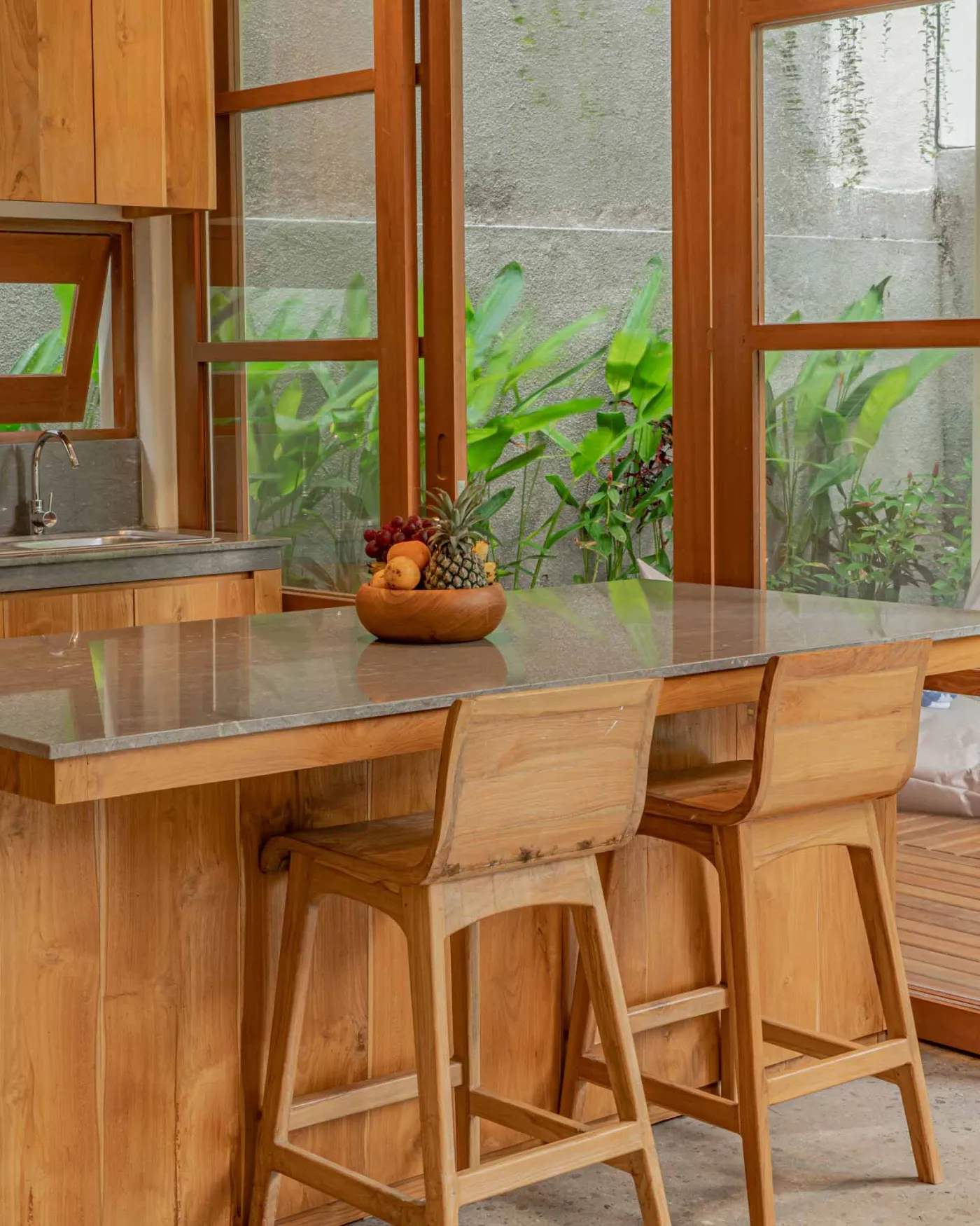 Spacious wooden kitchen and dining area in a tropical Balinese-style villa, featuring a set table with plates, glasses, vibrant floral centerpiece, and lush garden view through open sliding doors. - Bali Villas