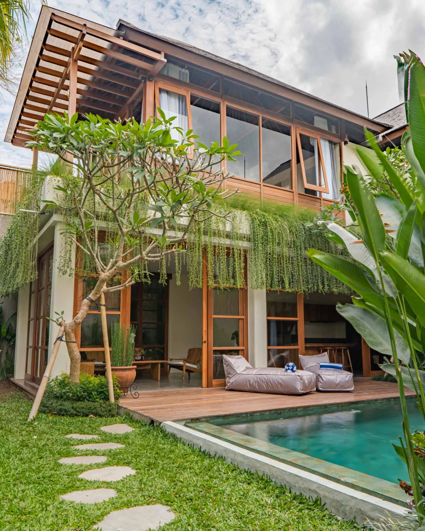Modern open-plan living and dining area in a tropical villa with wooden furniture, large sliding doors to poolside garden, and lush greenery. - Bali Villas