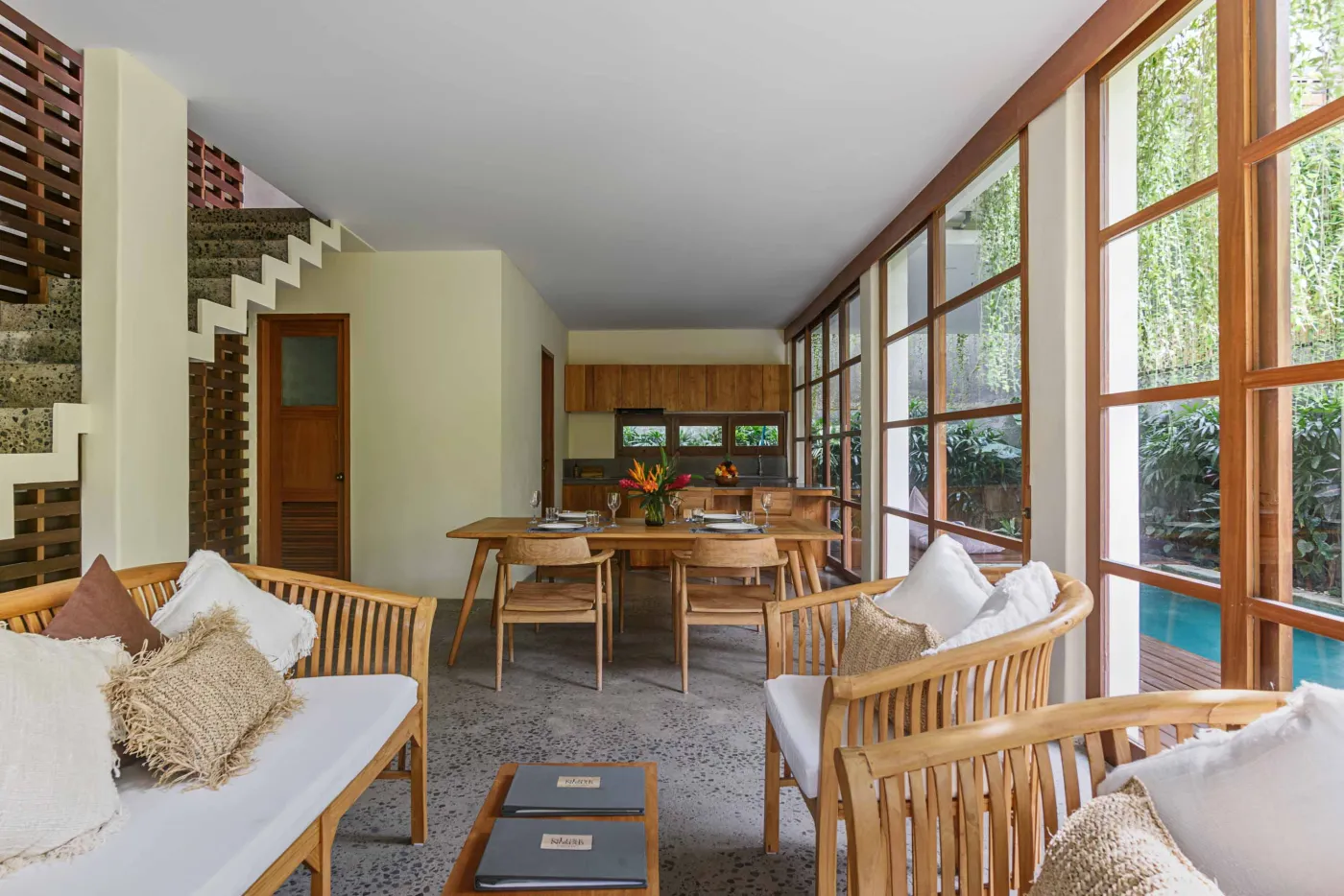 Modern open-plan living and dining area in a tropical villa with wooden furniture, large sliding doors to poolside garden, and lush greenery. - Bali Villas