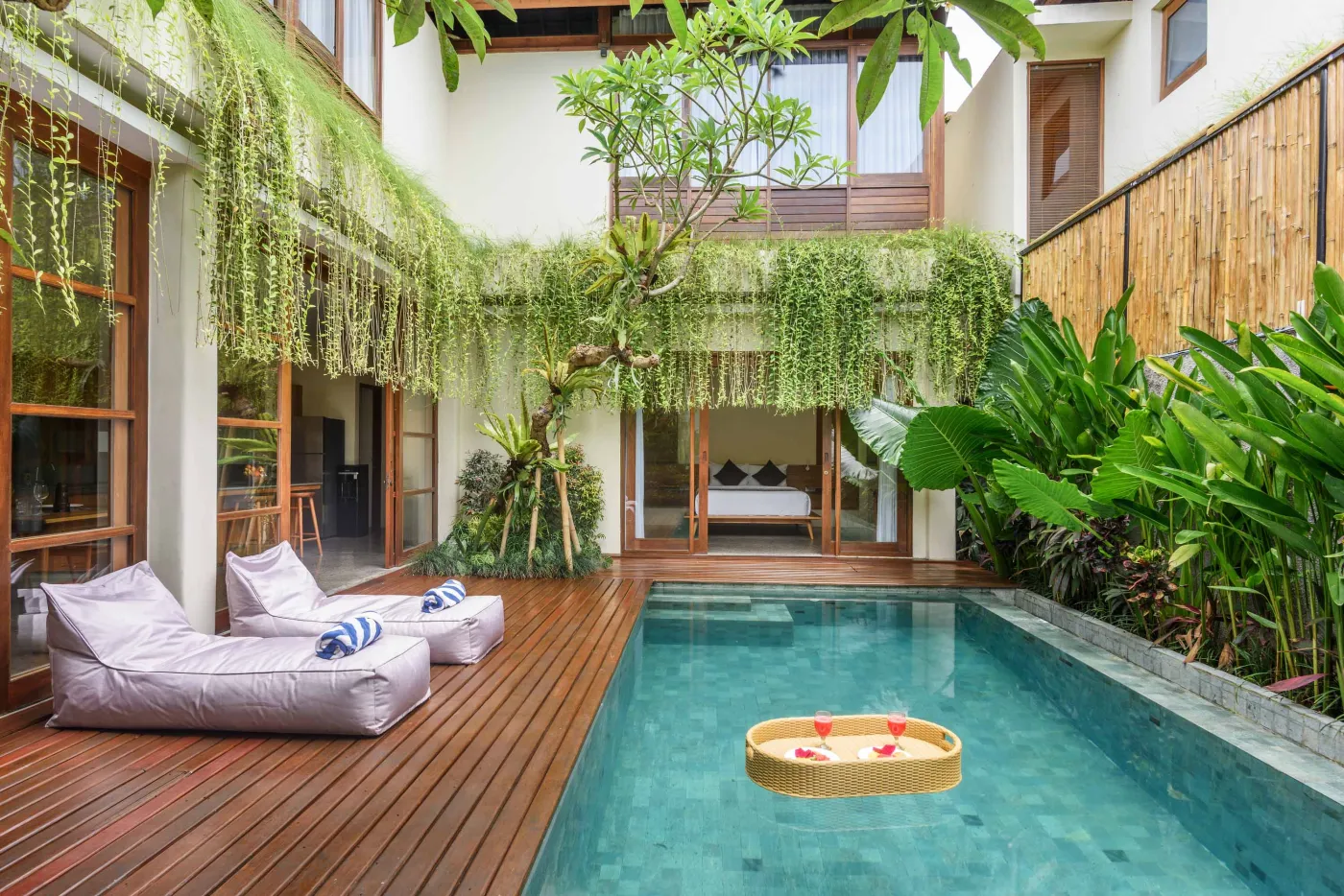 Modern tropical wooden kitchen with gray countertop island, rattan bar stools, potted plants, and sliding doors to a lush poolside view. - Bali Villas