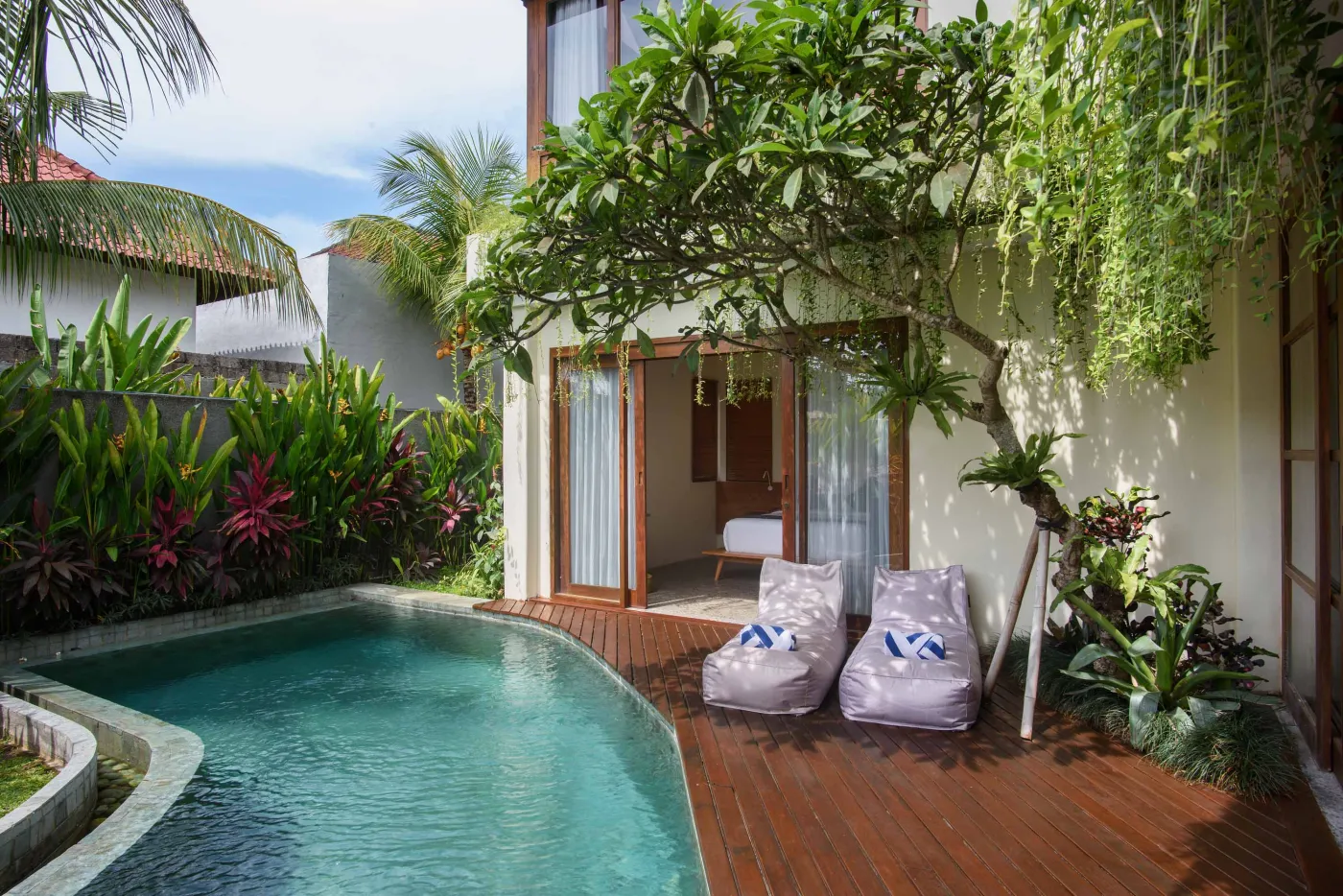 Modern tropical wooden kitchen with gray countertop island, rattan bar stools, potted plants, and sliding doors to a lush poolside view. - Bali Villas