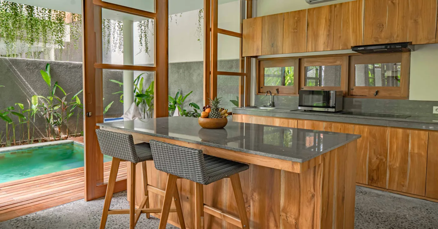 Modern tropical wooden kitchen with gray countertop island, rattan bar stools, potted plants, and sliding doors to a lush poolside view. - Bali Villas