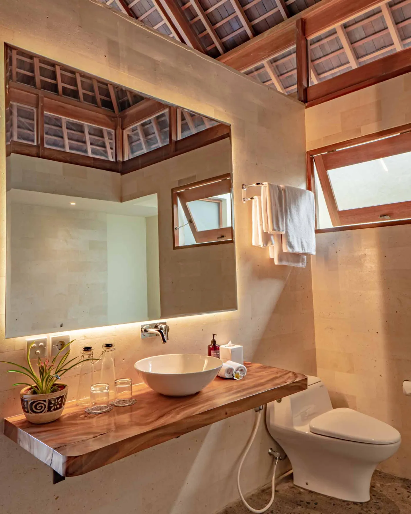 Modern tropical wooden kitchen with gray countertop island, rattan bar stools, potted plants, and sliding doors to a lush poolside view. - Bali Villas
