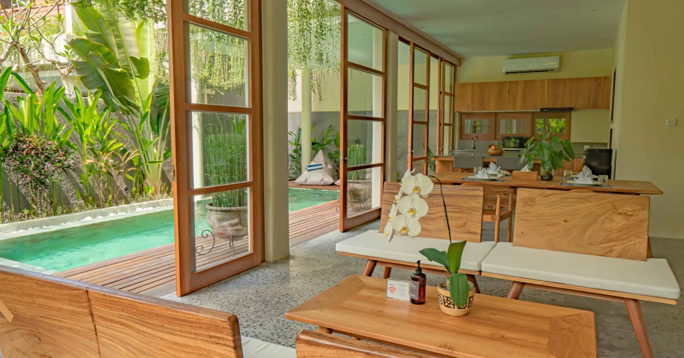 Modern tropical wooden kitchen with gray countertop island, rattan bar stools, potted plants, and sliding doors to a lush poolside view. - Bali Villas