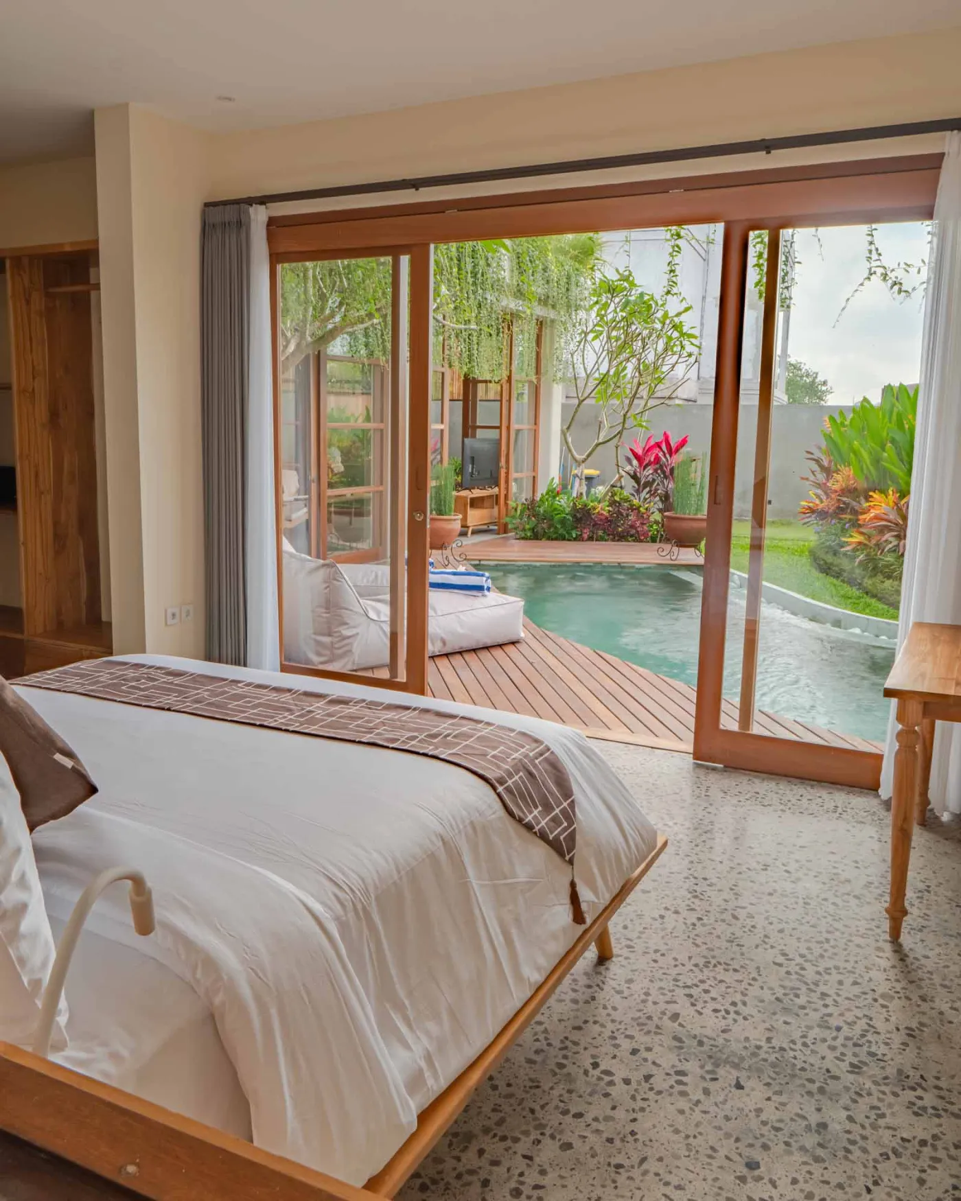 Modern tropical living room with wooden sofa, armchairs, low coffee table, potted orchid, wall-mounted TV, and open doors to lush garden. - Bali Villas