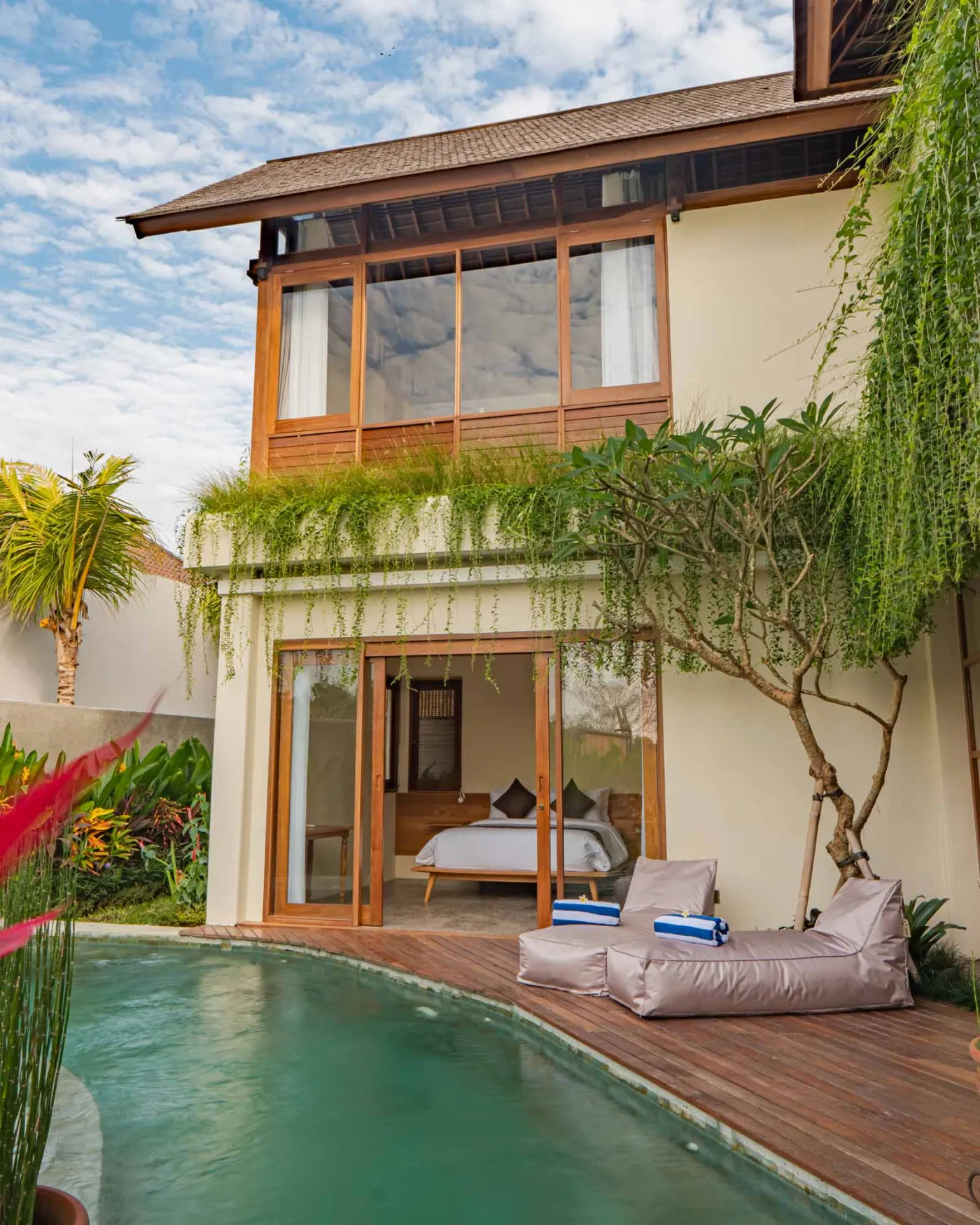 Modern tropical living room with wooden sofa, armchairs, low coffee table, potted orchid, wall-mounted TV, and open doors to lush garden. - Bali Villas