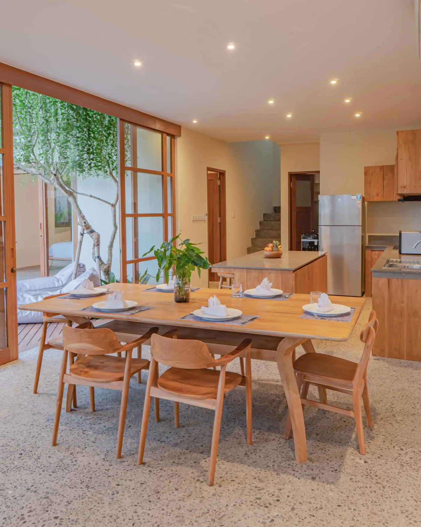 Modern tropical living room with wooden sofa, armchairs, low coffee table, potted orchid, wall-mounted TV, and open doors to lush garden. - Bali Villas