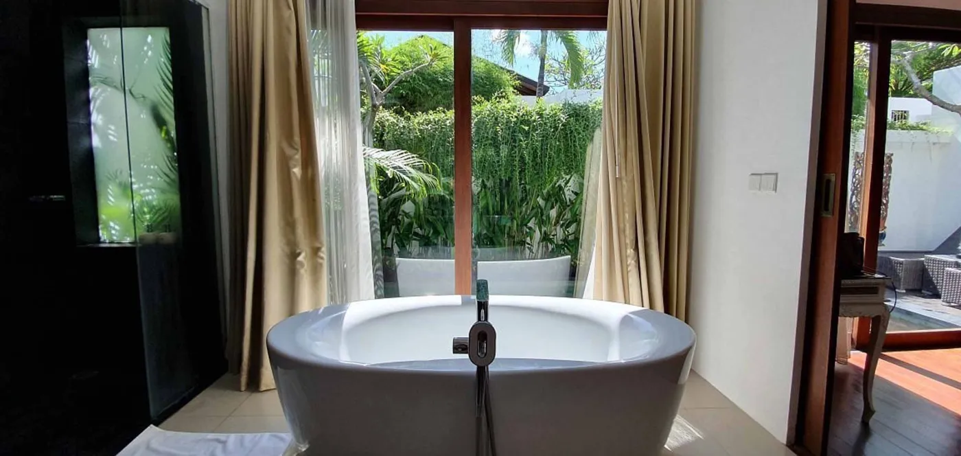 Bright modern Bali villa kitchen and dining area with wooden cabinets, rattan table topped with tropical fruits, ceiling fan, and sliding doors to lush garden. - Bali Villas