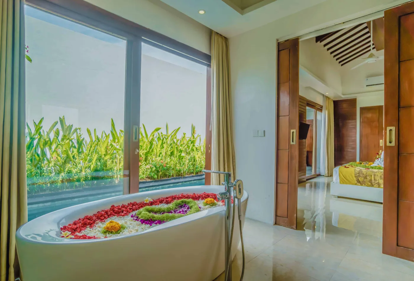 Bright modern Bali villa kitchen and dining area with wooden cabinets, rattan table topped with tropical fruits, ceiling fan, and sliding doors to lush garden. - Bali Villas