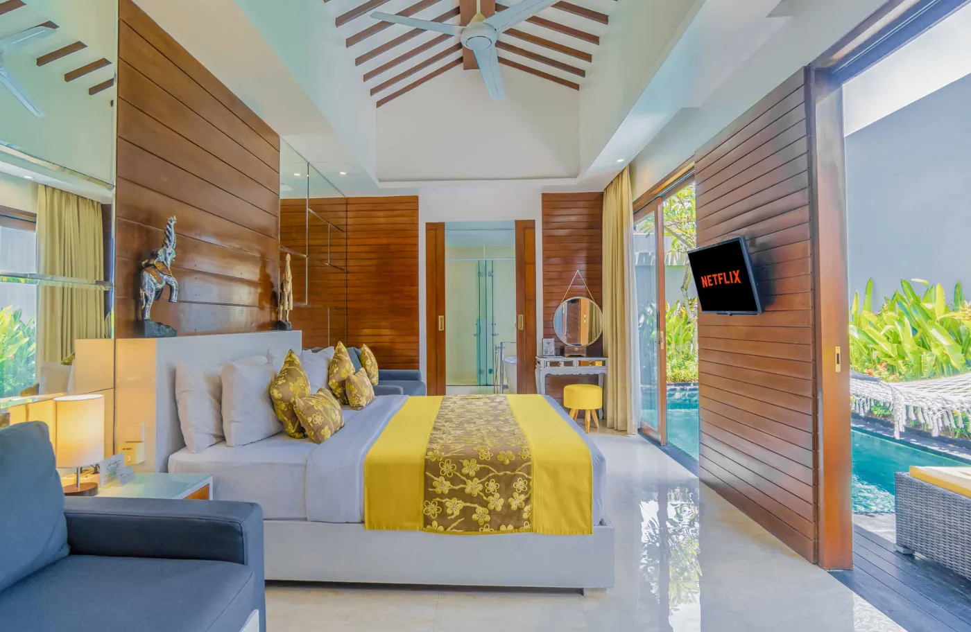 Bright modern Bali villa kitchen and dining area with wooden cabinets, rattan table topped with tropical fruits, ceiling fan, and sliding doors to lush garden. - Bali Villas