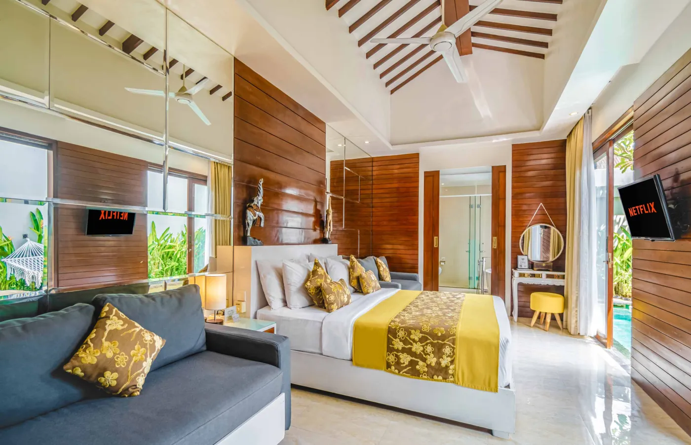 Bright modern Bali villa kitchen and dining area with wooden cabinets, rattan table topped with tropical fruits, ceiling fan, and sliding doors to lush garden. - Bali Villas