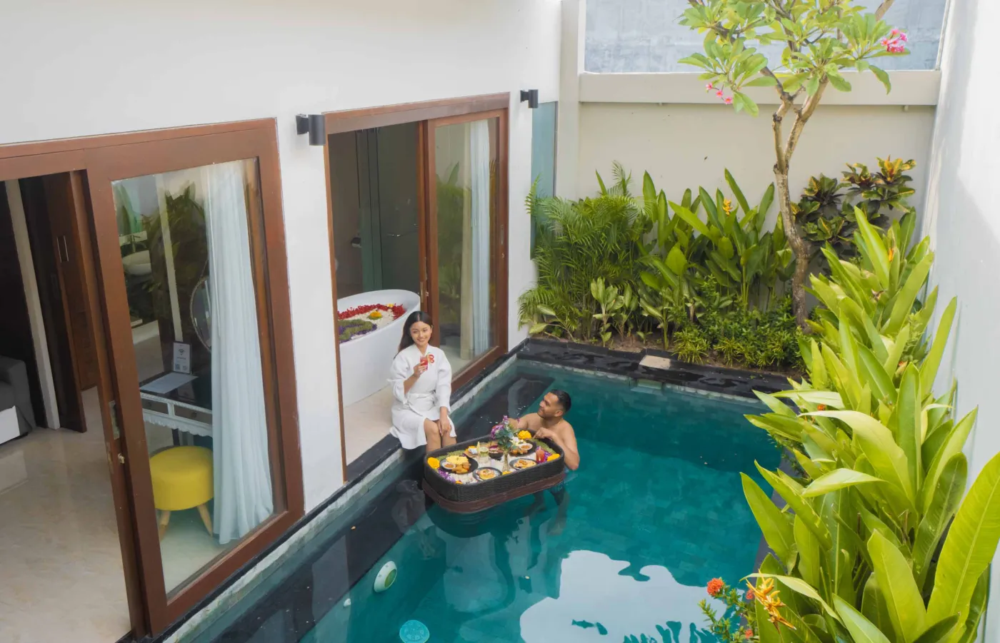 Bright modern Bali villa kitchen and dining area with wooden cabinets, rattan table topped with tropical fruits, ceiling fan, and sliding doors to lush garden. - Bali Villas