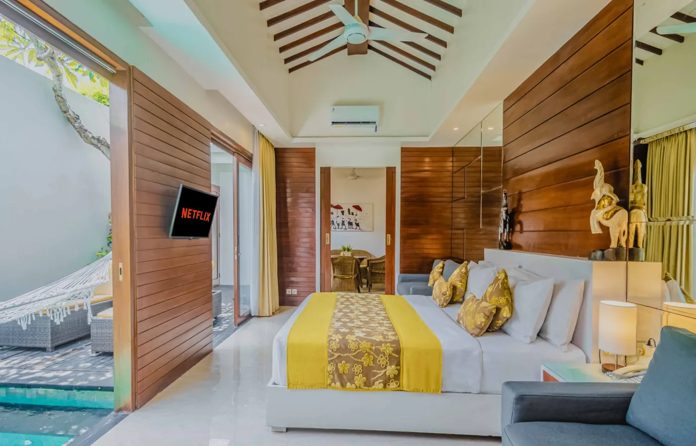Bright modern Bali villa kitchen and dining area with wooden cabinets, rattan table topped with tropical fruits, ceiling fan, and sliding doors to lush garden. - Bali Villas