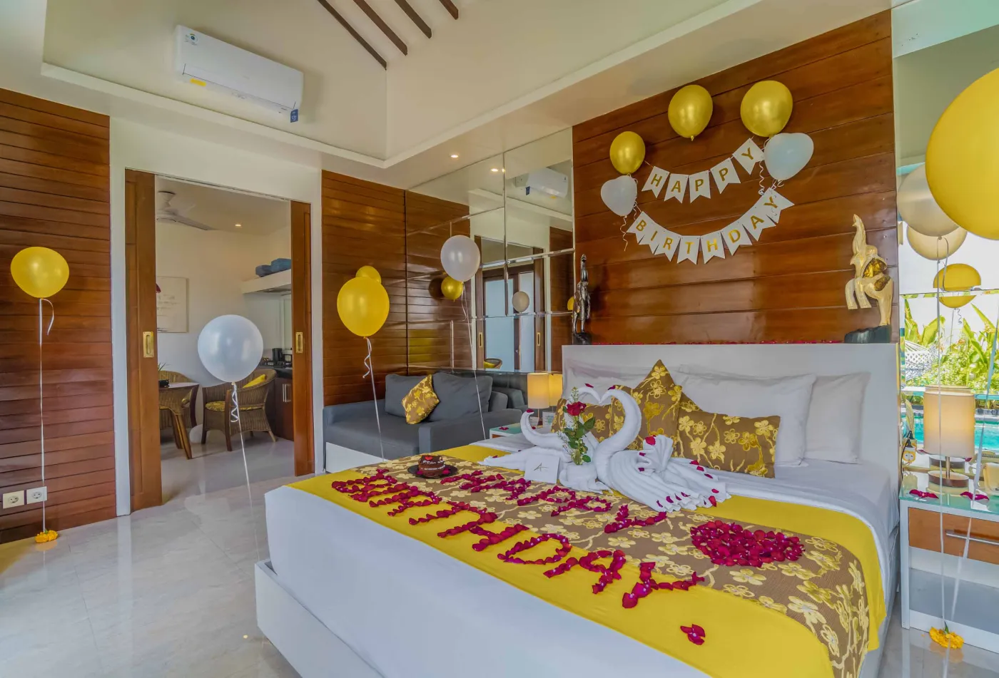 Bright modern Bali villa kitchen and dining area with wooden cabinets, rattan table topped with tropical fruits, ceiling fan, and sliding doors to lush garden. - Bali Villas