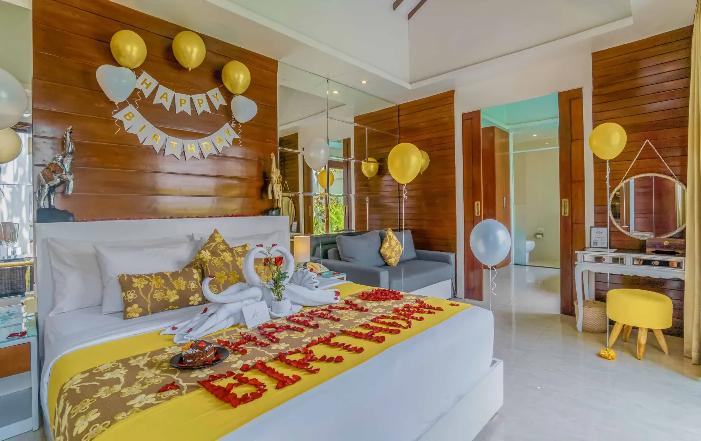 Bright modern Bali villa kitchen and dining area with wooden cabinets, rattan table topped with tropical fruits, ceiling fan, and sliding doors to lush garden. - Bali Villas