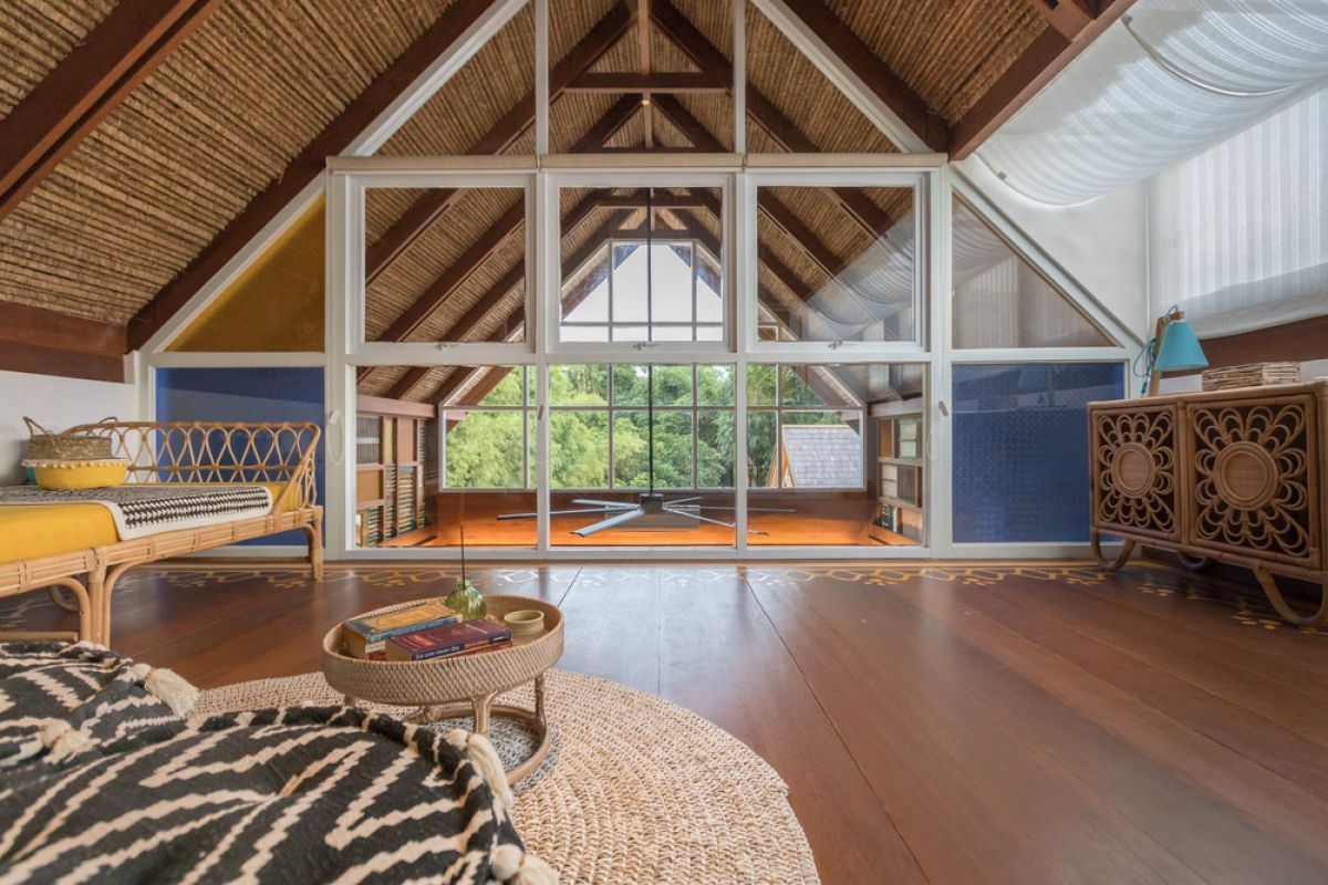 Spacious tropical Balinese villa interior with high wooden-beamed ceiling, large glass windows overlooking lush garden, plush sectional sofa, and modern kitchen bar. - Bali Villas