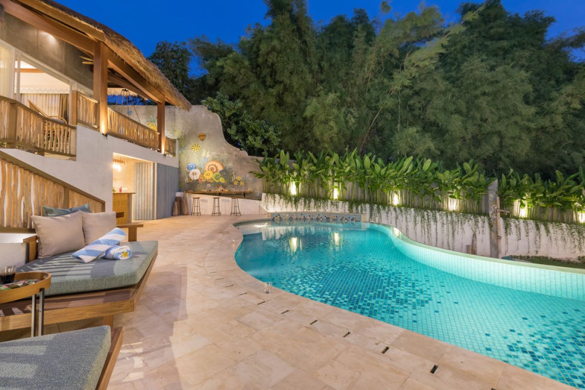 Spacious tropical Balinese villa interior with high wooden-beamed ceiling, large glass windows overlooking lush garden, plush sectional sofa, and modern kitchen bar. - Bali Villas