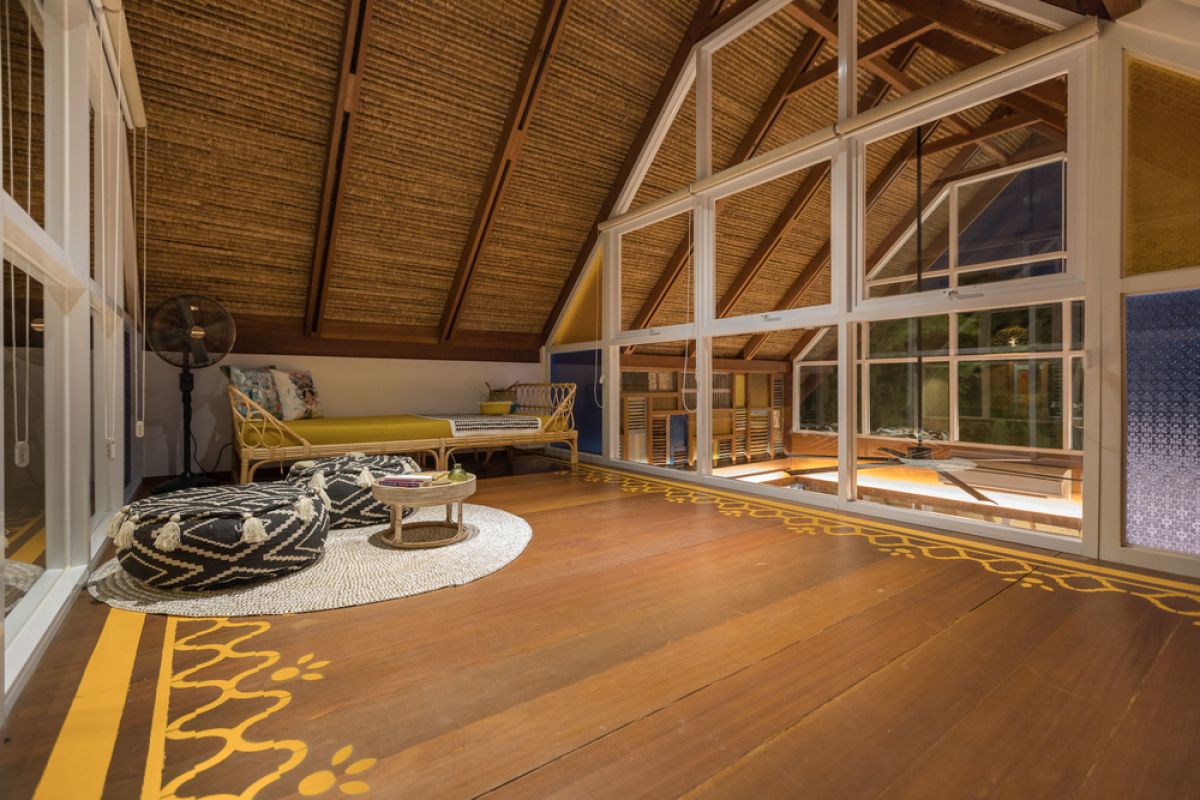 Spacious tropical Balinese villa interior with high wooden-beamed ceiling, large glass windows overlooking lush garden, plush sectional sofa, and modern kitchen bar. - Bali Villas