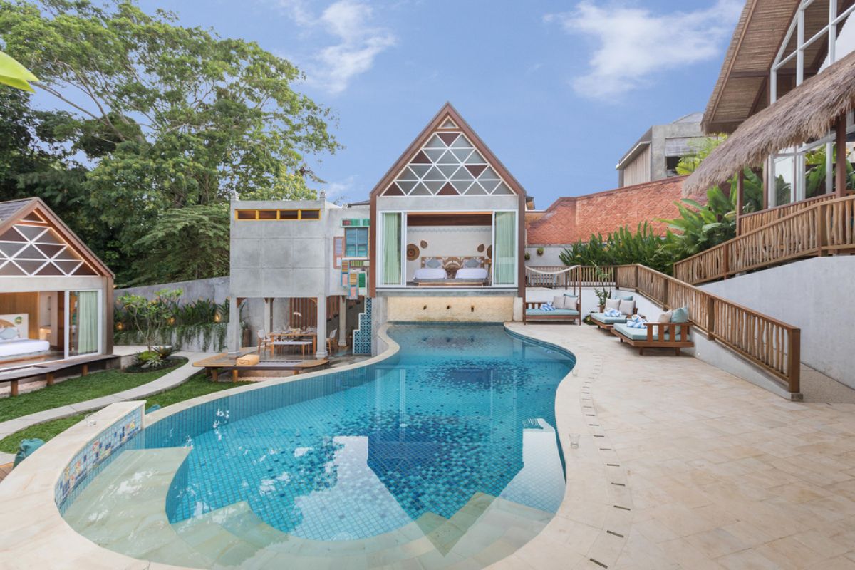 Spacious tropical Balinese villa interior with high wooden-beamed ceiling, large glass windows overlooking lush garden, plush sectional sofa, and modern kitchen bar. - Bali Villas