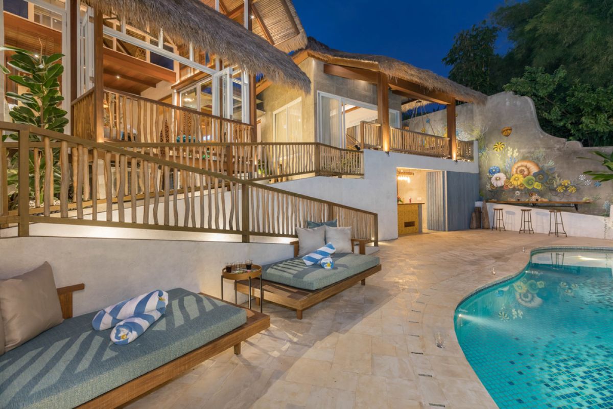 Spacious tropical Balinese villa interior with high wooden-beamed ceiling, large glass windows overlooking lush garden, plush sectional sofa, and modern kitchen bar. - Bali Villas