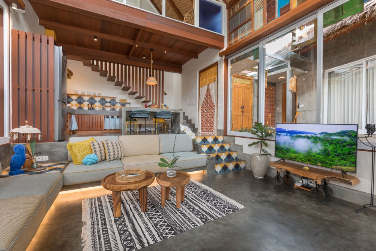 Spacious tropical Balinese villa interior with high wooden-beamed ceiling, large glass windows overlooking lush garden, plush sectional sofa, and modern kitchen bar. - Bali Villas