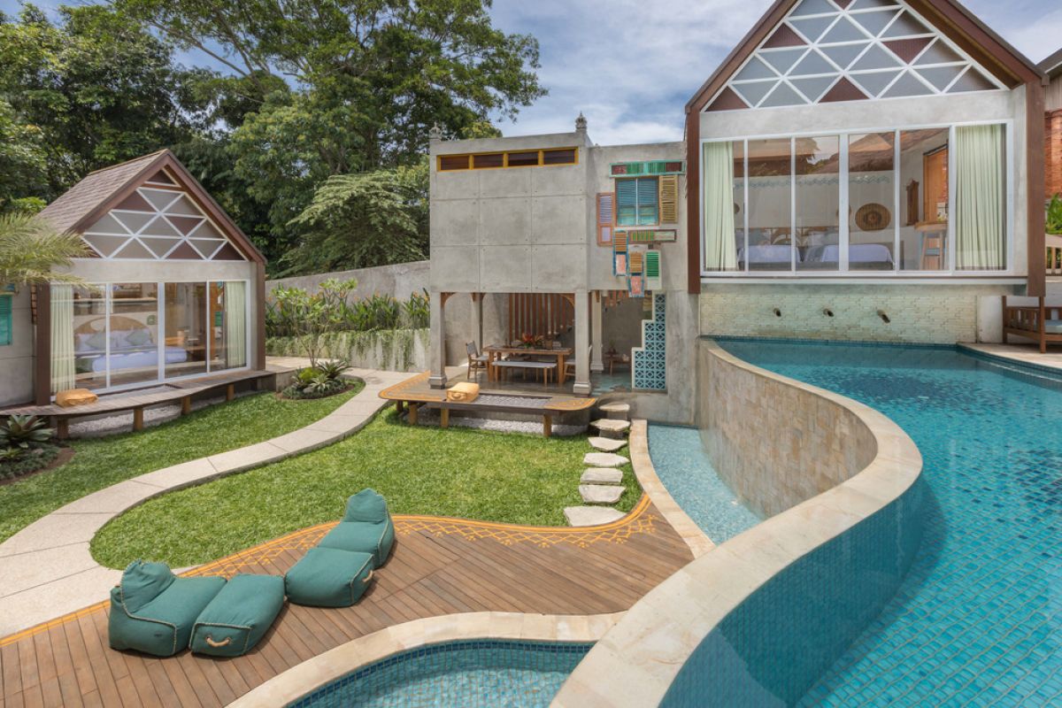 Spacious tropical Balinese villa interior with high wooden-beamed ceiling, large glass windows overlooking lush garden, plush sectional sofa, and modern kitchen bar. - Bali Villas