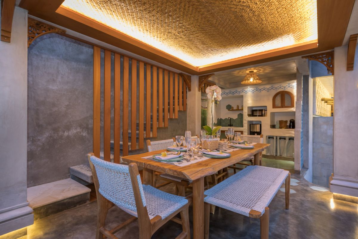 Spacious tropical Balinese villa interior with high wooden-beamed ceiling, large glass windows overlooking lush garden, plush sectional sofa, and modern kitchen bar. - Bali Villas
